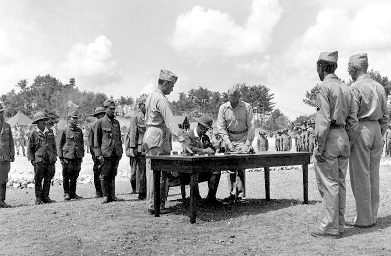 TENTH ARMY HEADQUARTERS, Okinawa, September 7, 1945: Lieutenant General Toshiro Nomi, representing Japanese forces in the Sakishima Islands, signs the surrender document. Standing beside him are Colonel Philip Bethune, of the G-2 Section, and Major General Frank Merrill, Tenth Army chief of staff. Standing opposite the table at right are MIS Nisei Robert H. Oda and Lieutenant General Joseph W. Stilwell, commanding general, Tenth Army. (U.S. Navy photo)